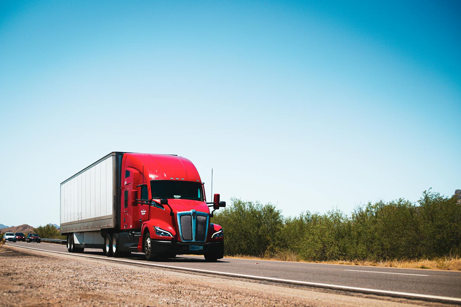 Vibrant red semi-trailer truck driving on an open highway under a clear blue sky.